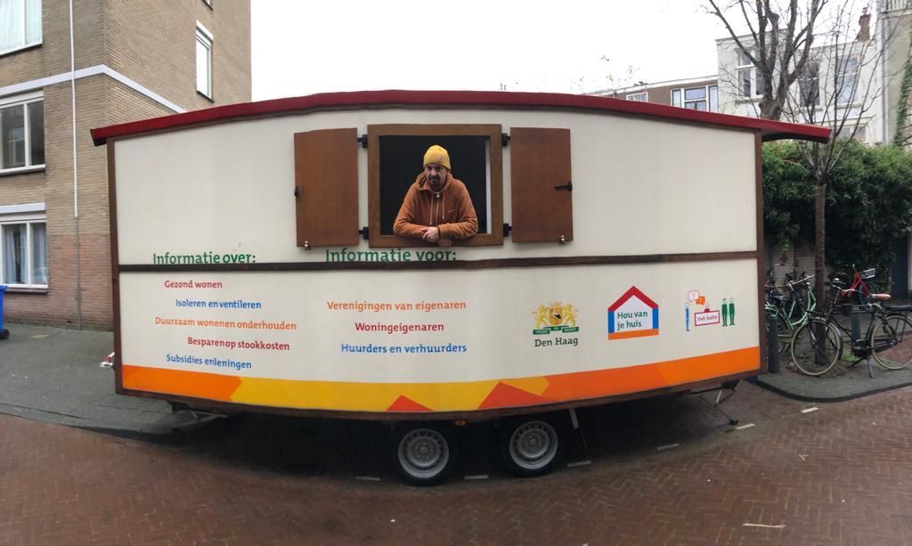 Iemand kijkt uit het raam van een stacaravan. De stacaravan is wit, met een bruin dak en een kleurrijk logo.