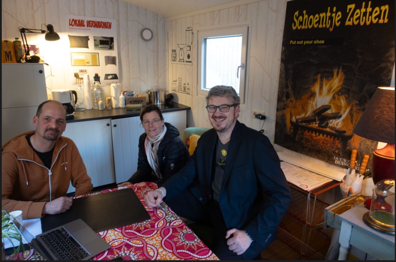 Drie mensen zitten aan een tafel. Interieurfoto, vol met allerlei spullen. Eén persoon leunt naar een laptop.