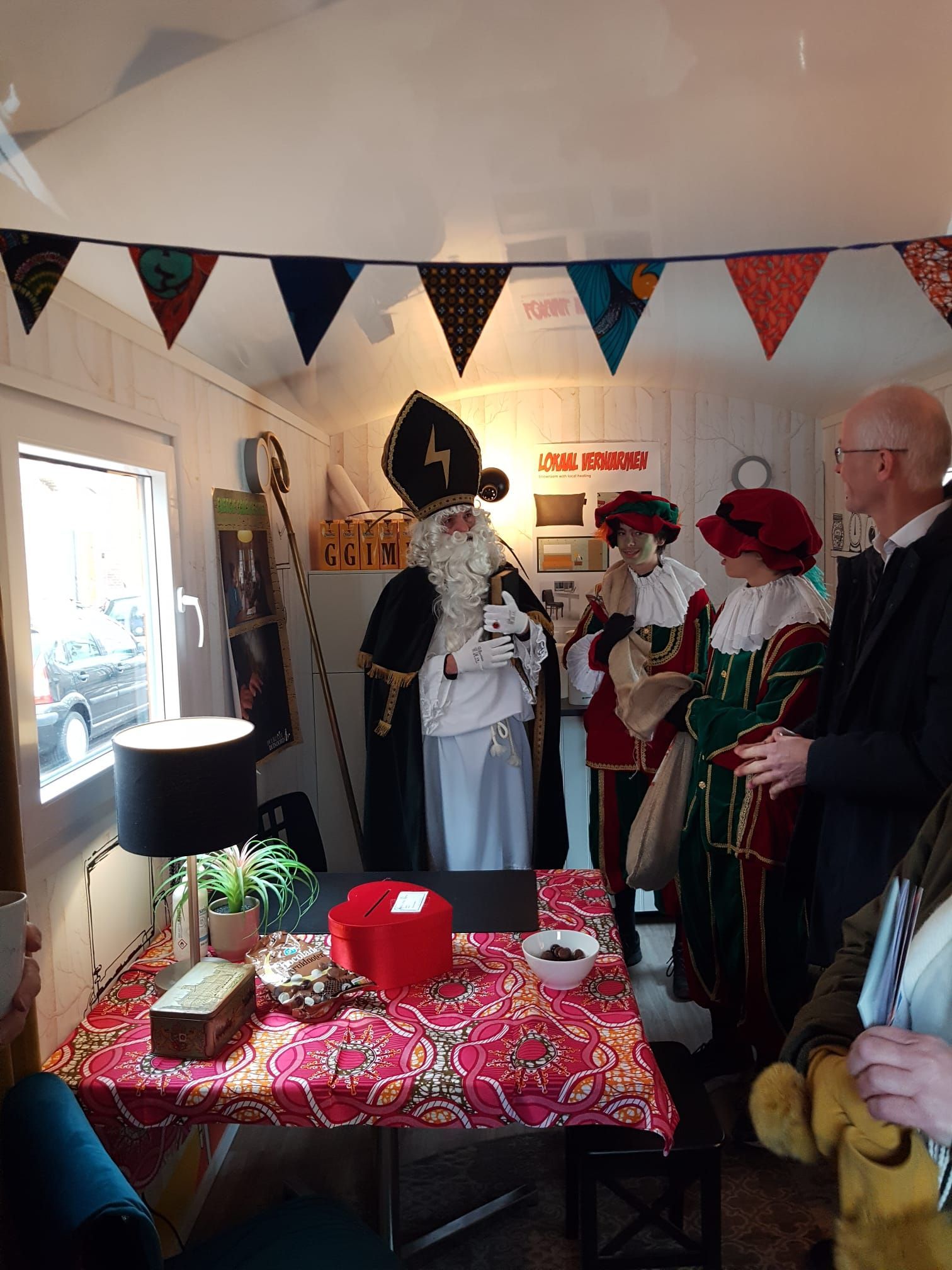 Sinterklaas (Sint Nicolaas) in rijkversierde gewaden en hoed, omringd door helpers, aan een versierde tafel binnenshuis.