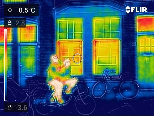 Thermische opname van mensen op een fiets voor een gebouw. ​​De afbeelding toont warmte in gele en oranje tinten.