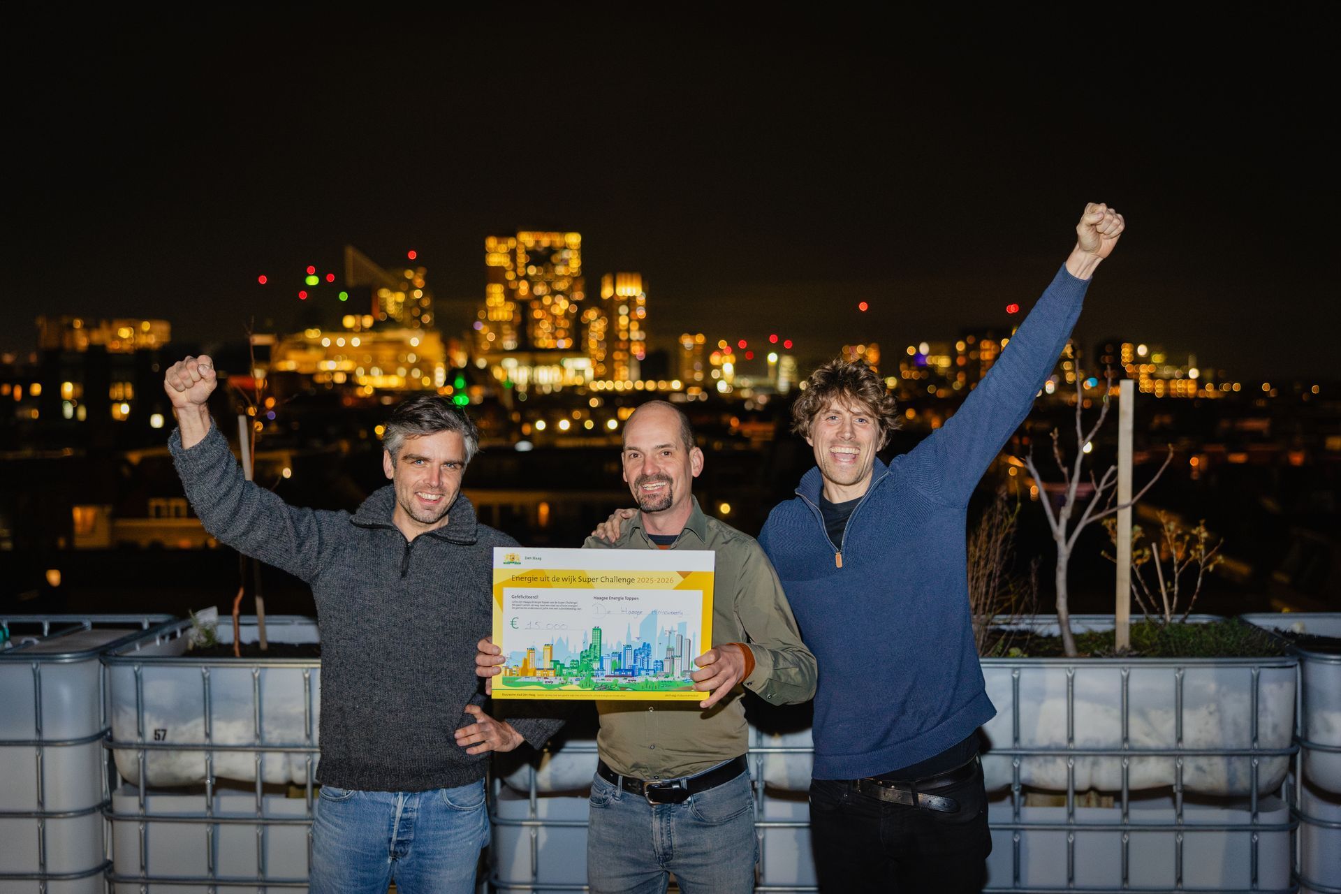 Drie mensen staan ​​'s nachts op een dak, lachend en met een ingelijste foto met een stadsgezicht op de achtergrond.