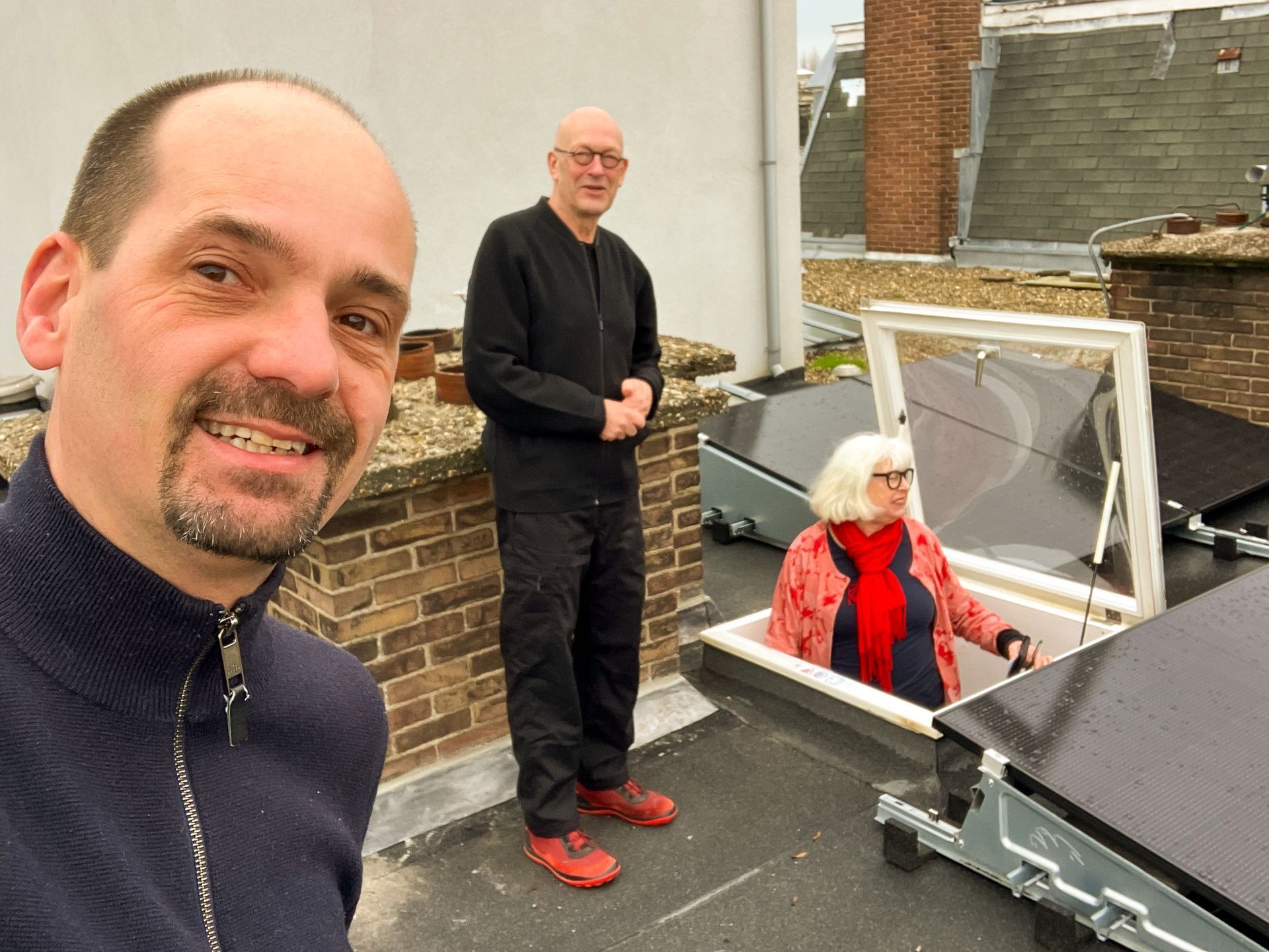 Drie mensen op een dakterras naast een open dakraam, waarvan er één een selfie maakt.