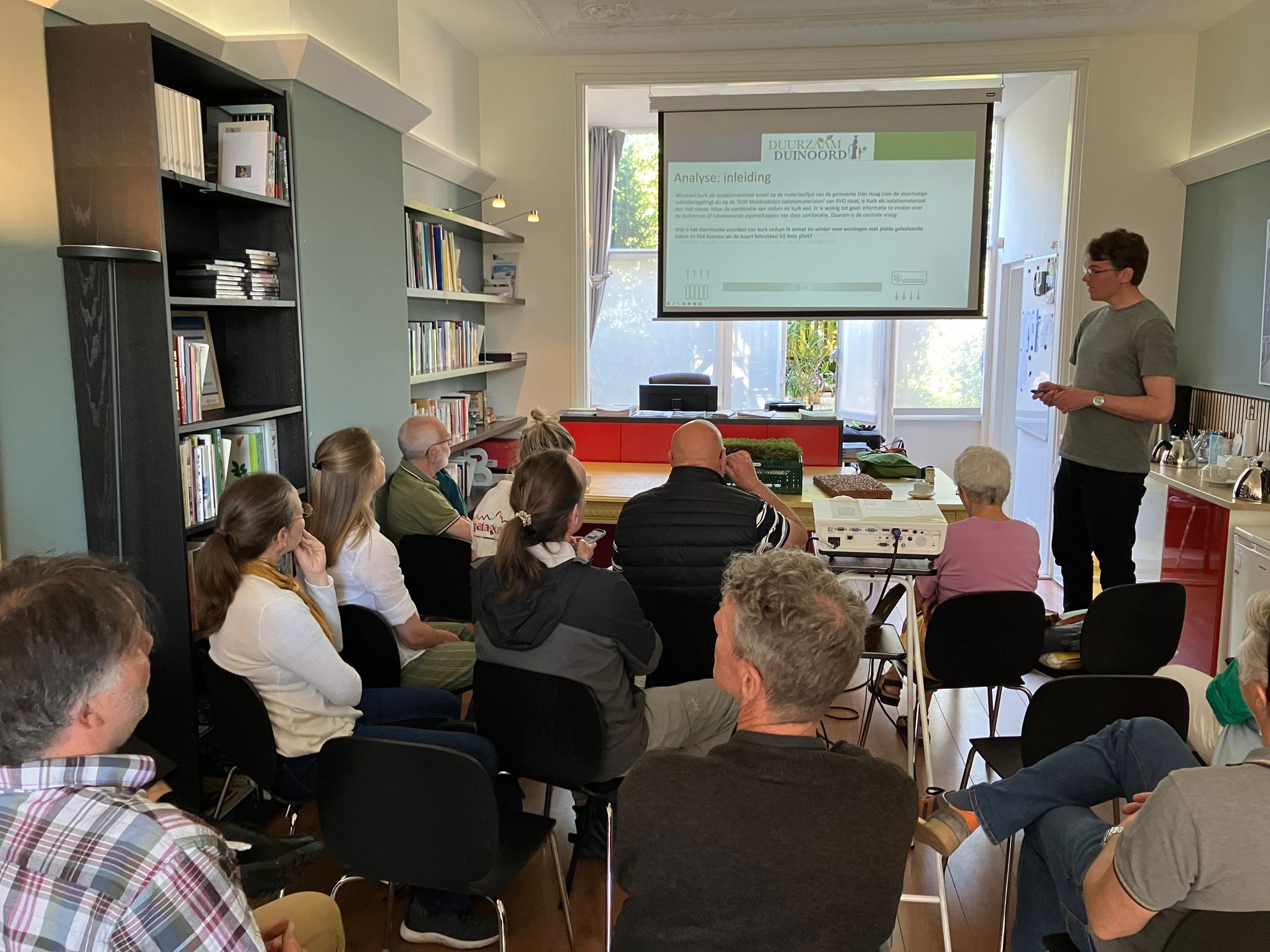 Het publiek zit in een klaslokaal dat is ingericht als een woonkamer en kijkt naar een geprojecteerde presentatie.