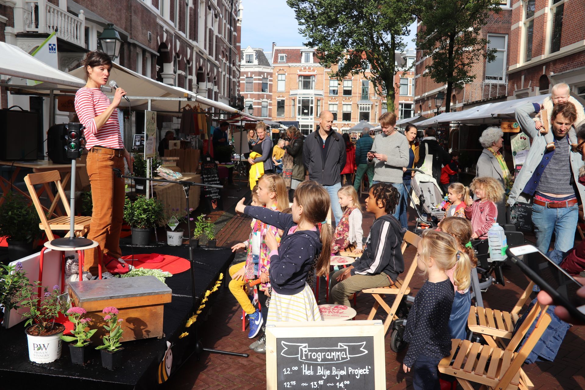 Een drukke openluchtmarkt met winkelend publiek, kraampjes, bloemen en een artiest op een klein podium.