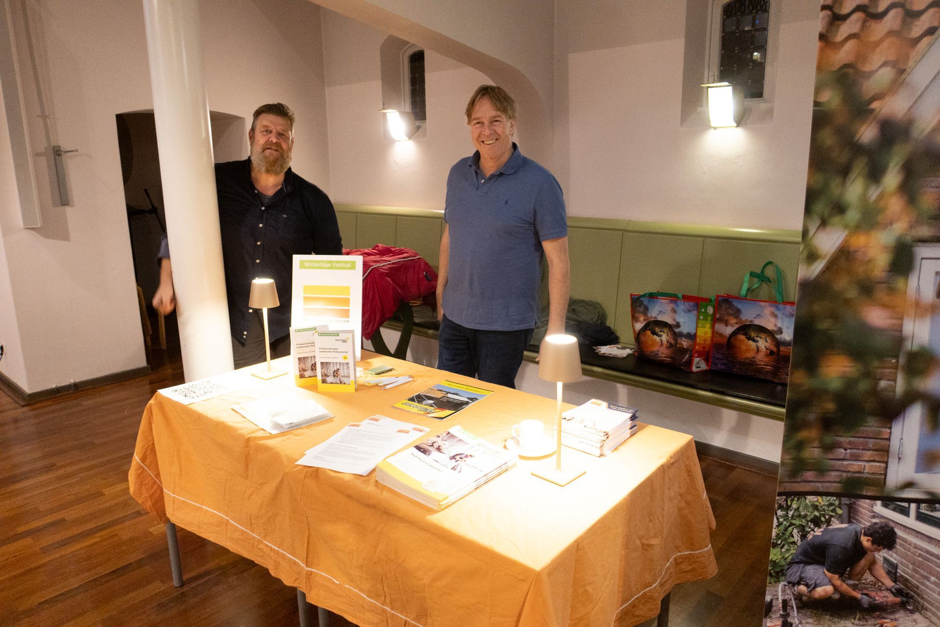 Twee mannen staan ​​achter een tafel met brochures en geschenken, waarschijnlijk bij een evenement in een gebouw.