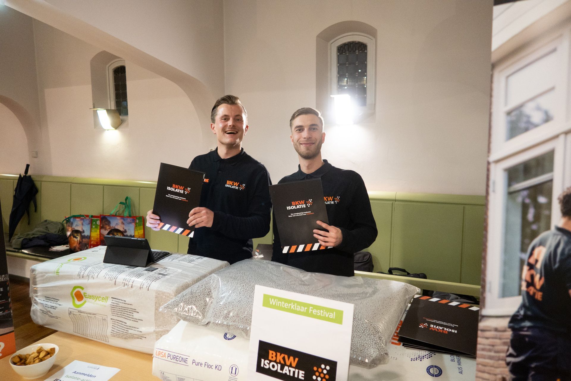 Twee mannen staan ​​bij een promotietafel en houden brochures vast. Op de tafel liggen artikelen die in plastic zijn afgedekt. ​​De ruimte is fel verlicht.