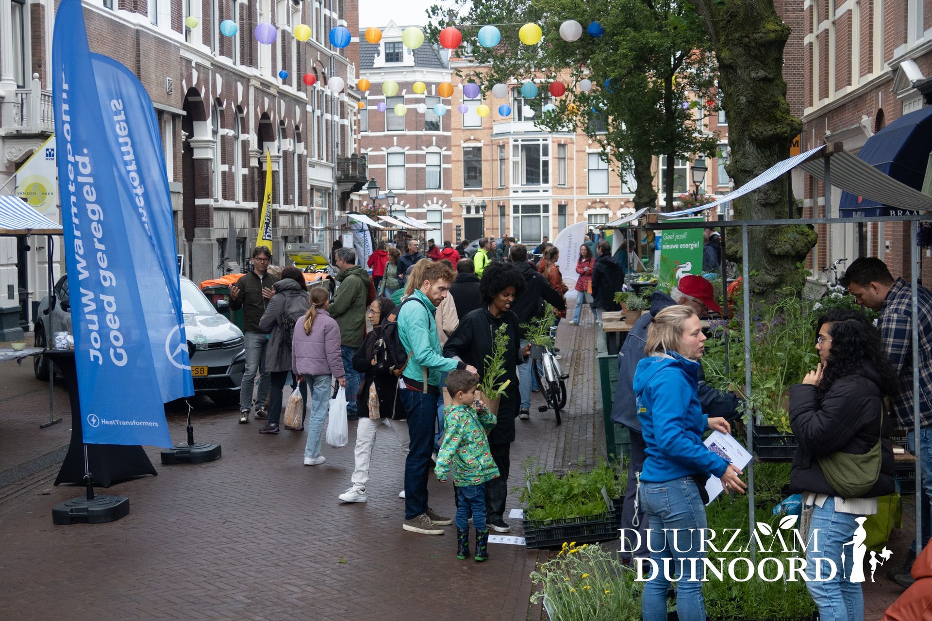 Een drukke straatmarkt met mensen die langs de kraampjes slenteren onder kleurrijke hangende versieringen in een stadscentrum.