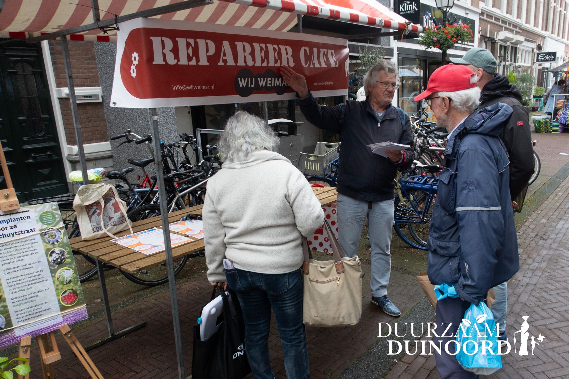 Mensen die rondkijken bij een cafékraam op een buitenmarkt, met fietsen en een rood bord met de tekst 