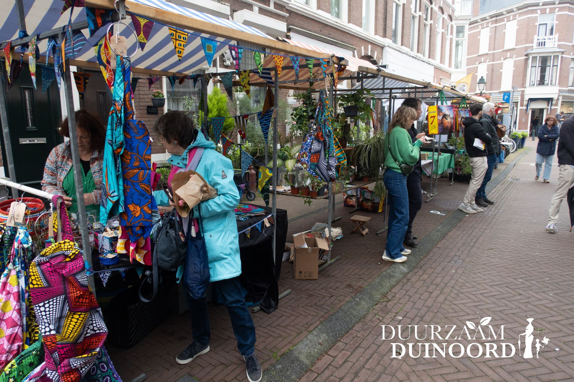 Marktkraampjes in de openlucht met kleurrijke hangende waren en winkelend publiek dat langs de straat slentert.