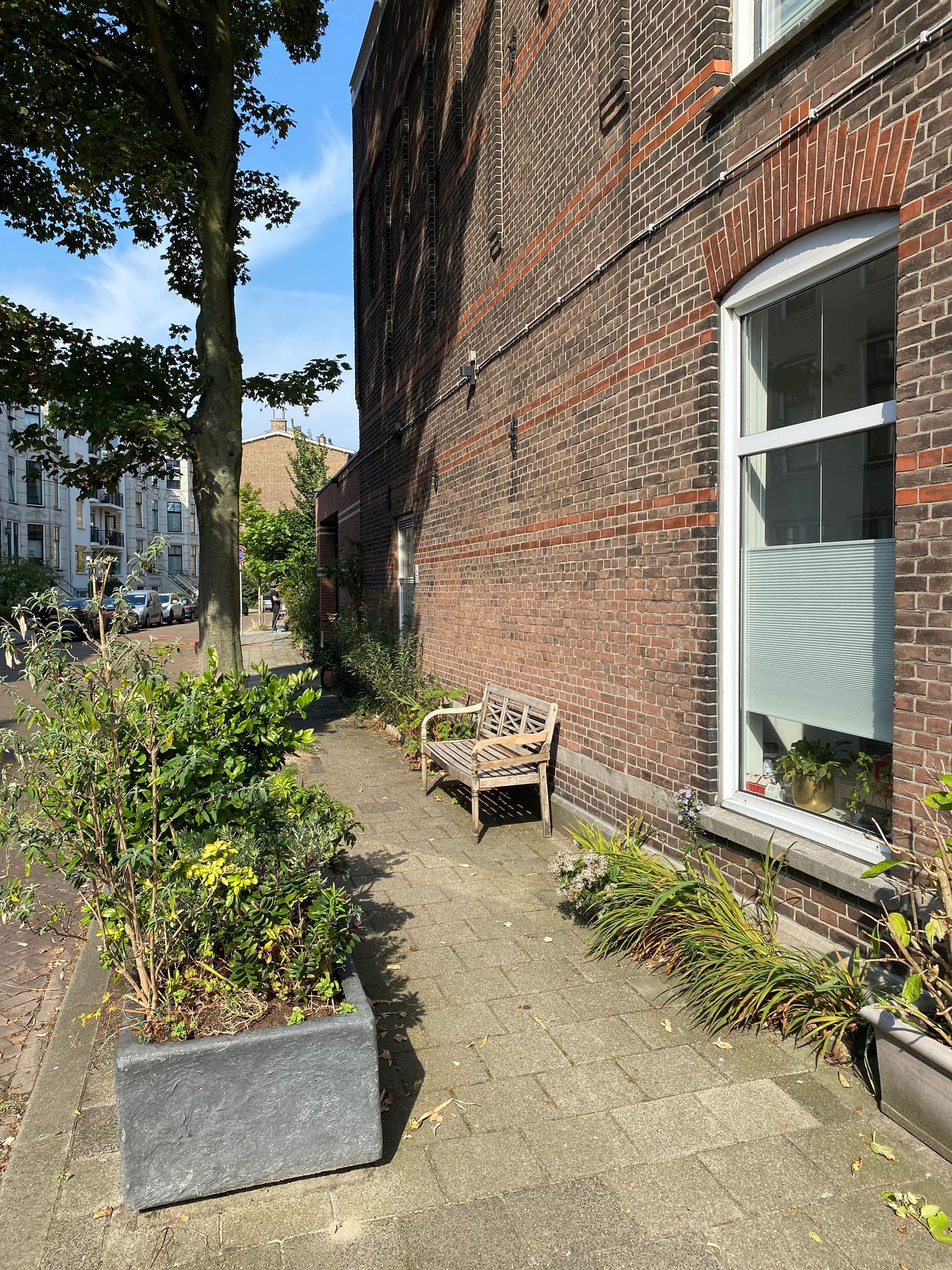Een bakstenen gevel met een klein bankje op een wandelpad omzoomd met struiken en een boom.