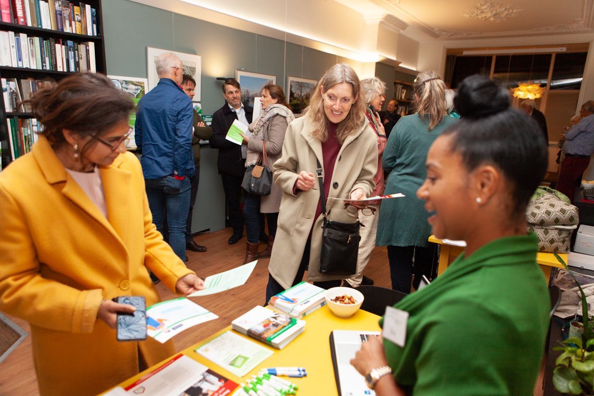 Mensen op een evenement, die met elkaar in gesprek zijn aan een tafel met brochures. Boekenkasten en een deuropening zijn zichtbaar.
