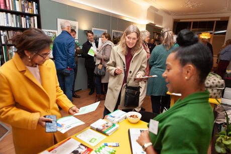 Mensen op een evenement, die met elkaar in gesprek zijn aan een tafel met brochures. Boekenkasten en een deuropening zijn zichtbaar.