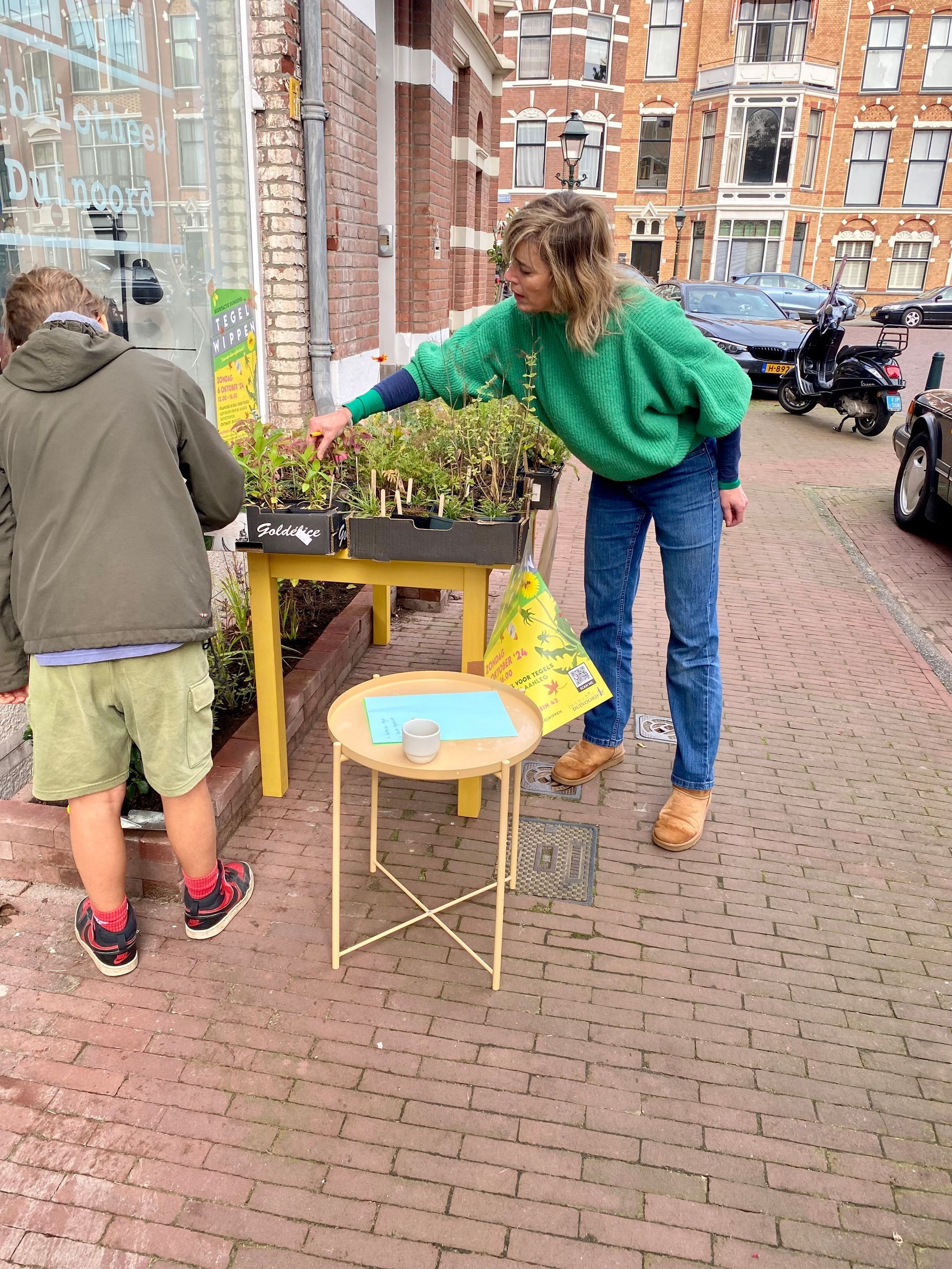 Iemand verkoopt planten op de stoep; een klant bekijkt ze. Gele tafel en een klein krukje. Bakstenen straat.