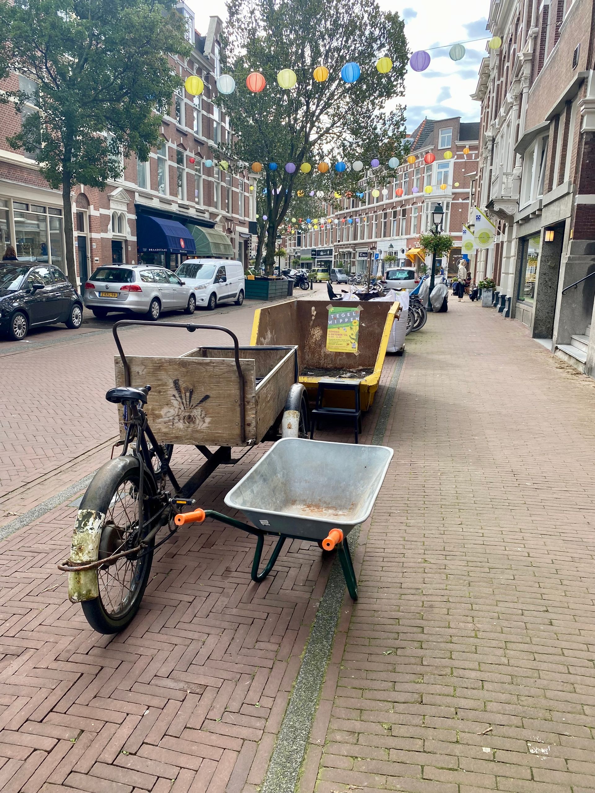 Een vintage fiets die drie containers trekt op een bakstenen straat met gebouwen en lichtslingers erboven.