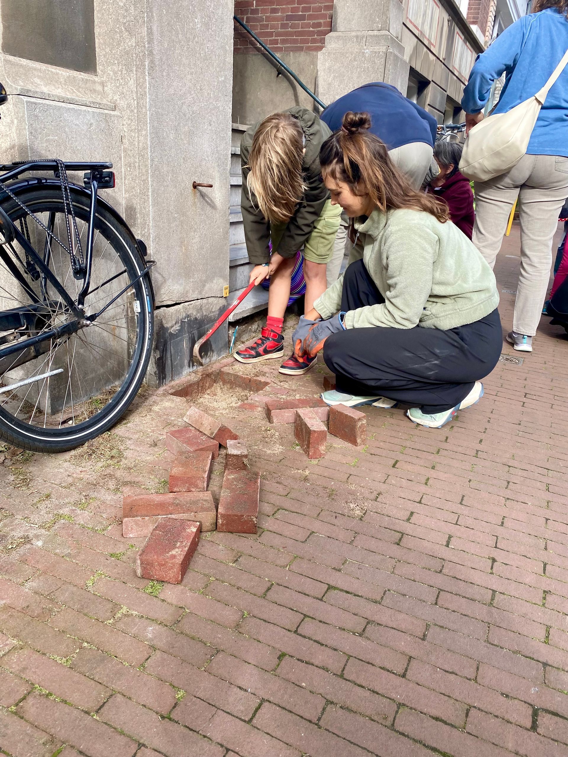 Mensen leggen stenen op een stoep bij een gebouw met een fiets links geparkeerd.
