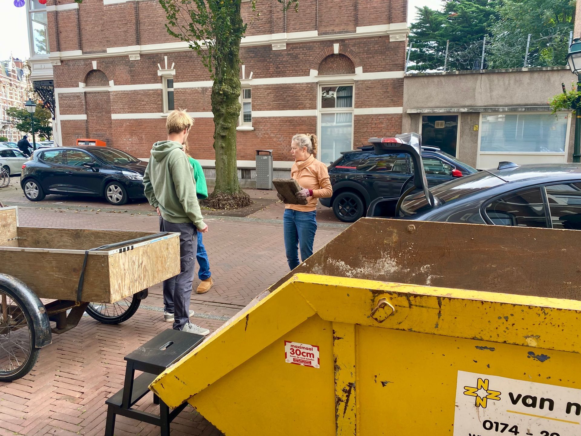 Mensen bij een gele afvalcontainer en een houten kar. Eén persoon houdt een klembord vast. Auto's staan ​​geparkeerd op straat.