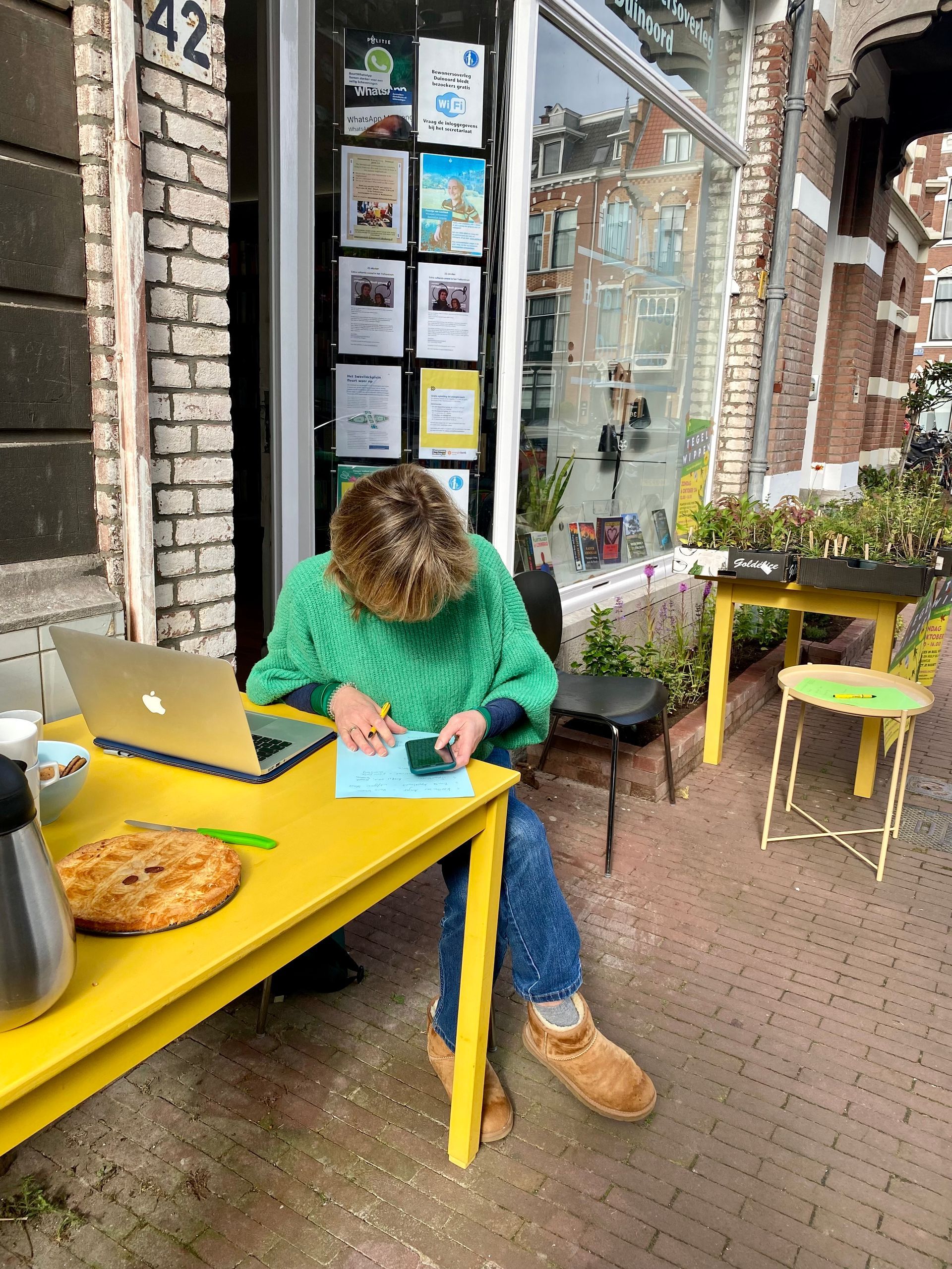 Een persoon werkt op een laptop aan een gele tafel buiten een winkel.