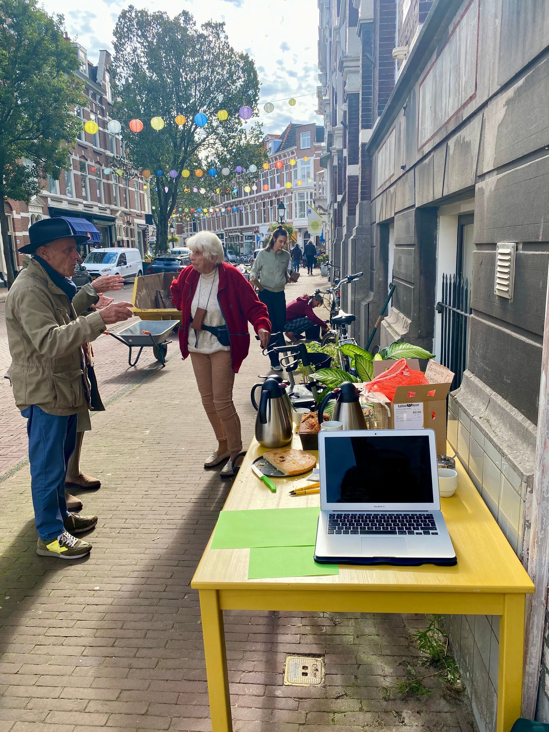 Mensen aan een tafel met eten, planten en een laptop op een stadsstoep.