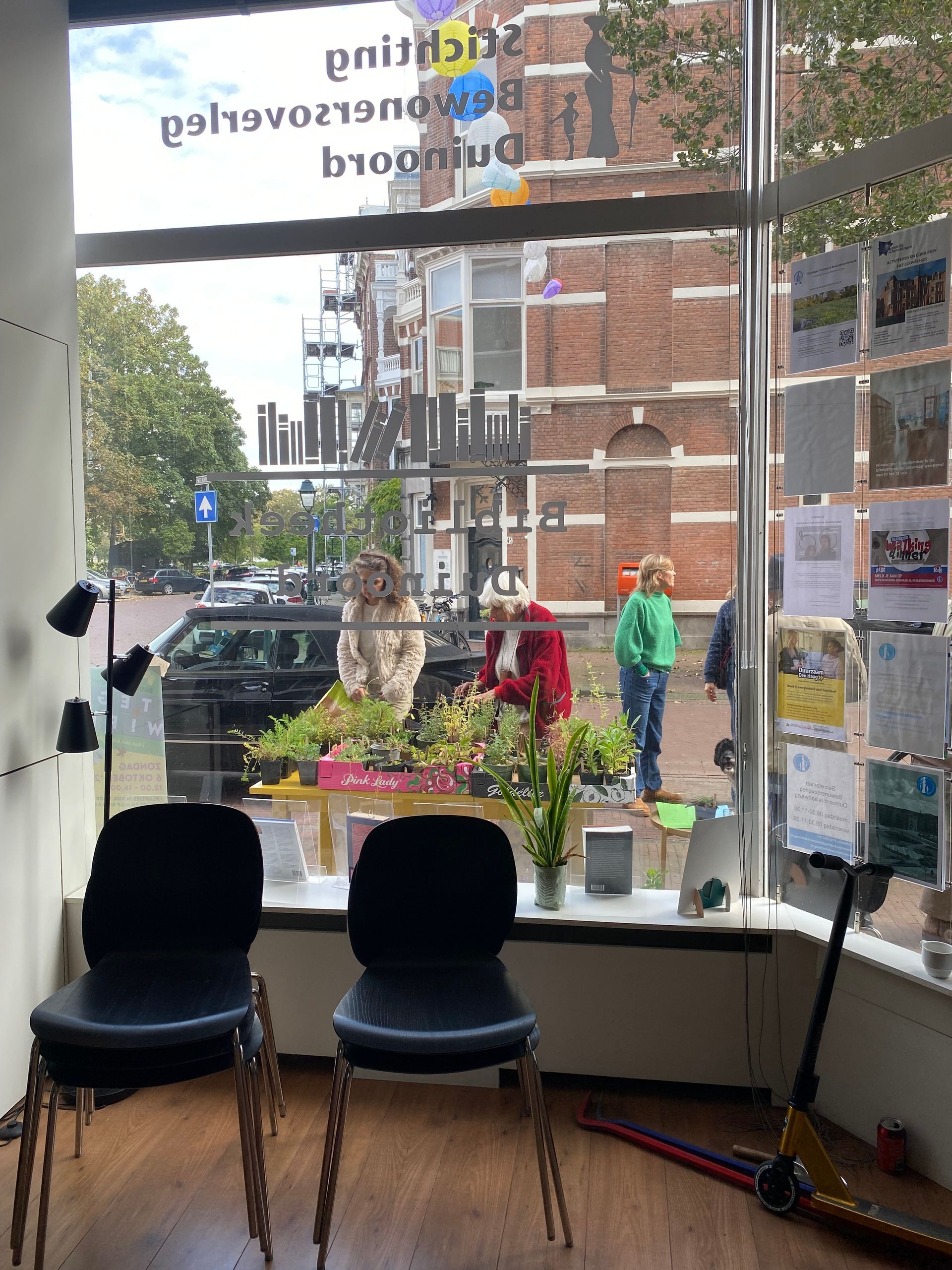 Twee zwarte stoelen bij een raam. Buiten: een persoon bij bloemen, voetgangers en een gebouw.