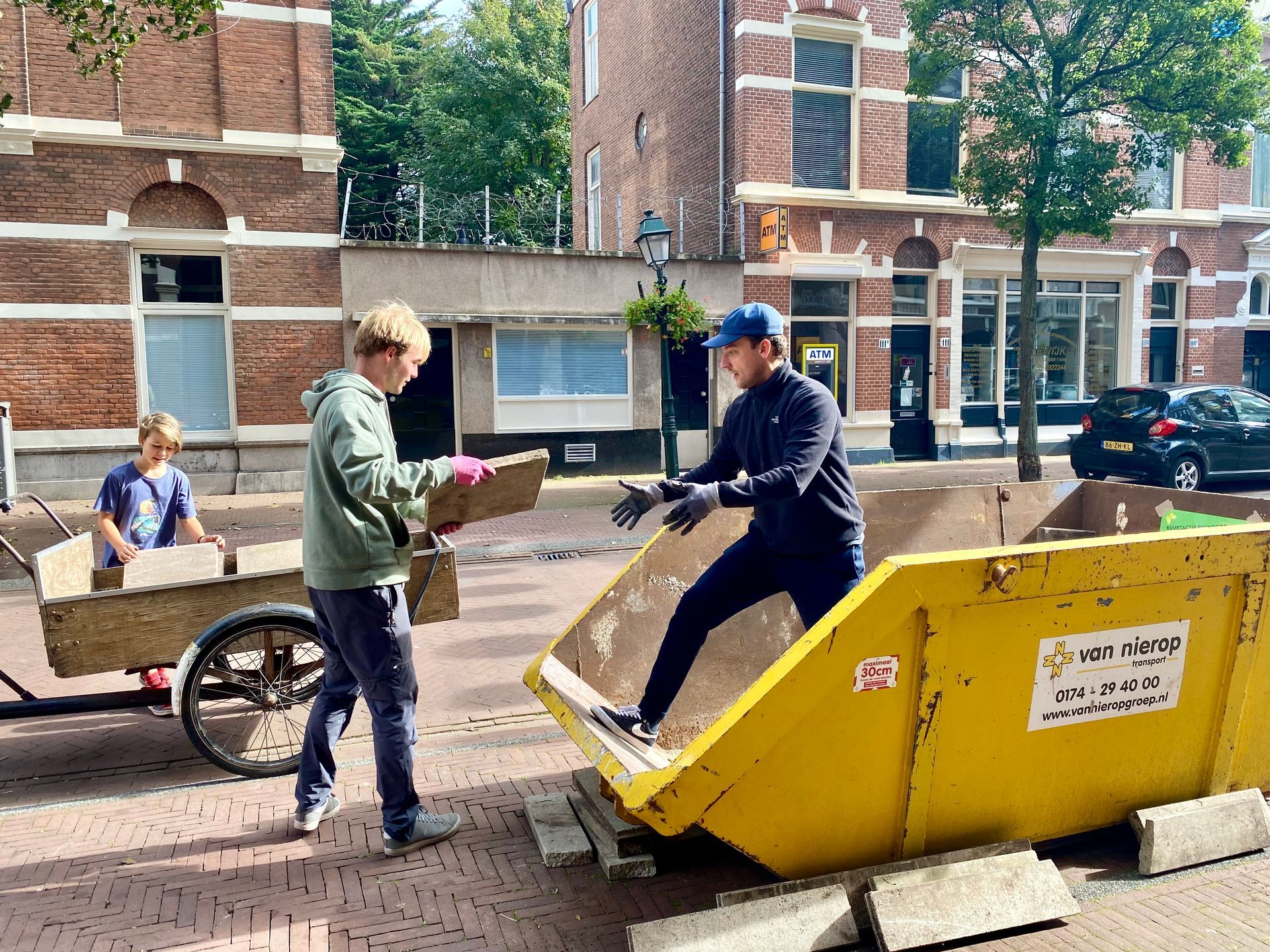 Drie mensen laden bakstenen in een gele afvalcontainer in een straat met bakstenen gebouwen.