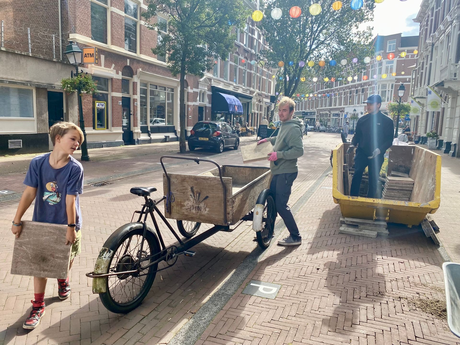 Een jongen draagt ​​een plank, een ander trekt een bakfiets voort, twee mensen zitten in een gele kar op een stadsstraat.