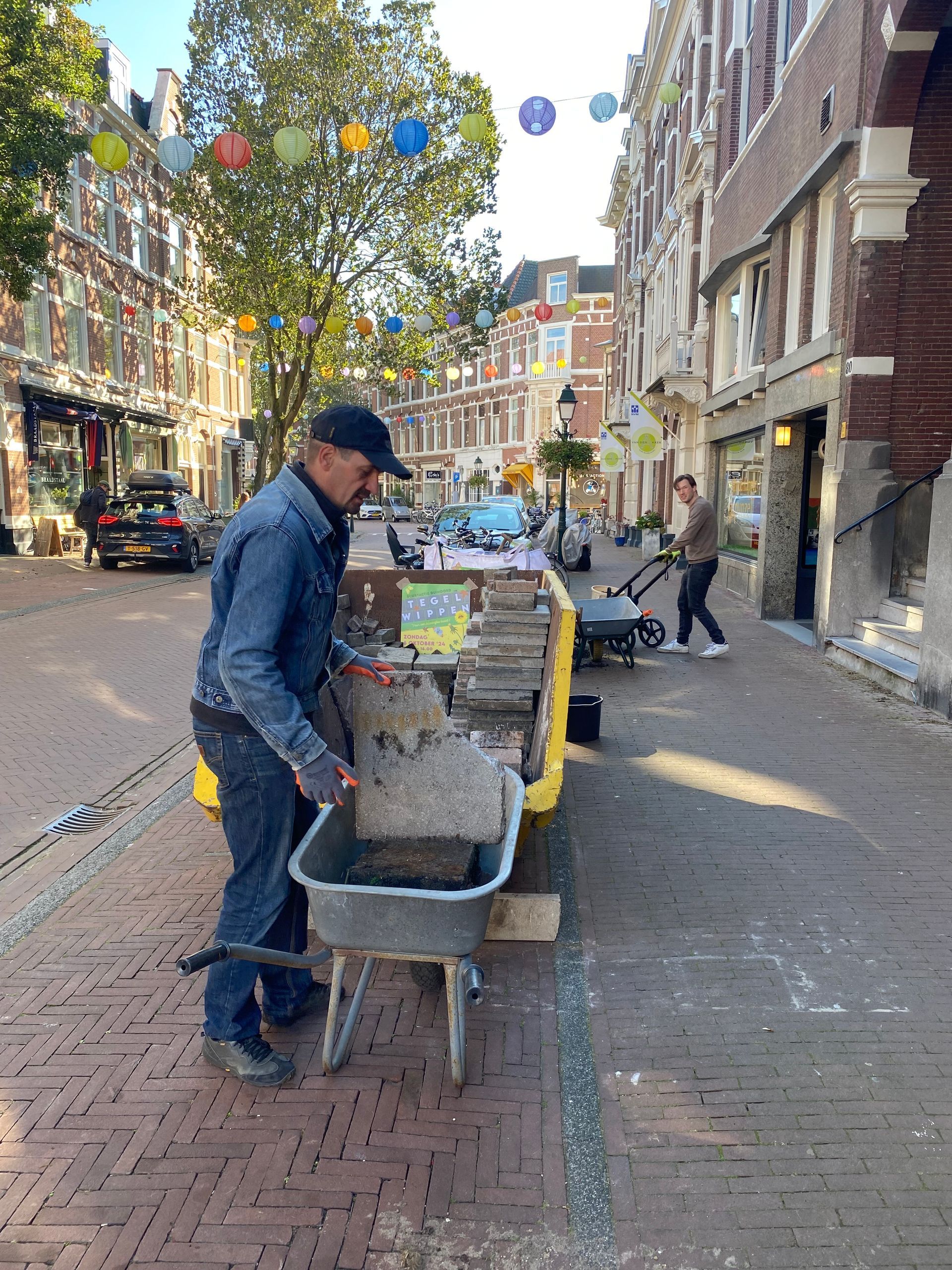 Een man gebruikt een kruiwagen om puin te verplaatsen op een bakstenen straat. Iemand anders gebruikt een bladblazer. Langs de straat staan ​​gebouwen.