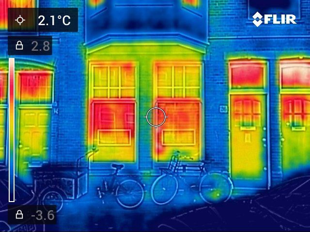 Thermische opname van een gebouwgevel die warmteverlies laat zien. Warme kleuren accentueren ramen en deuren; een fiets is zichtbaar.