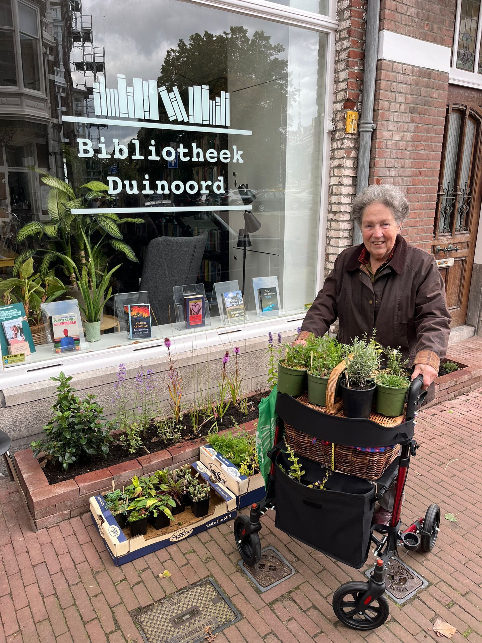 Een vrouw met een rollator staat voor een etalage met planten in een bibliotheek.