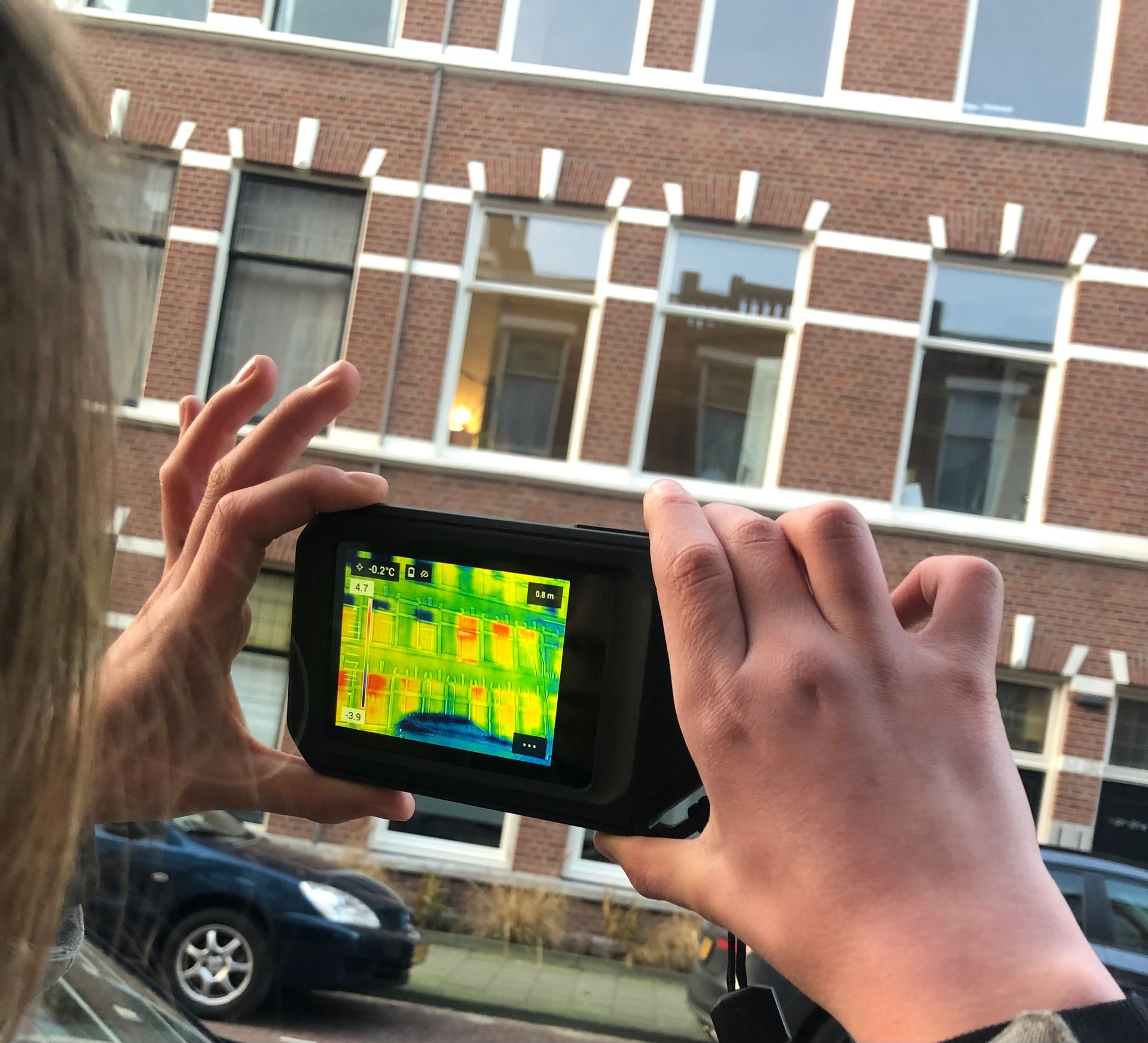 Een persoon gebruikt een warmtebeeldcamera om de gevel van een gebouw te scannen, waardoor warmteverschillen in kleurcodes op een display worden weergegeven.