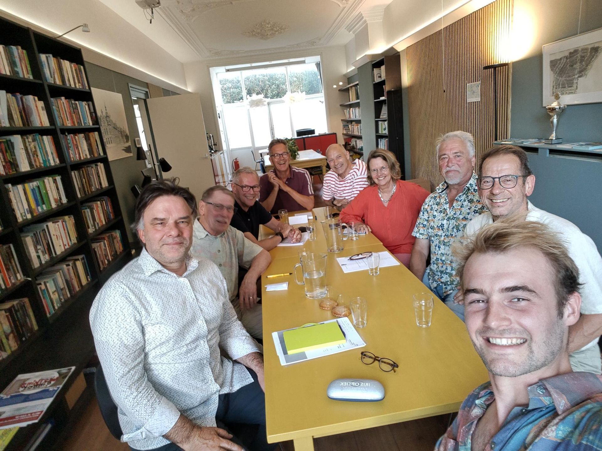 Een groepje mensen zit rond een gele tafel in een knusse kamer en lacht voor een selfie.