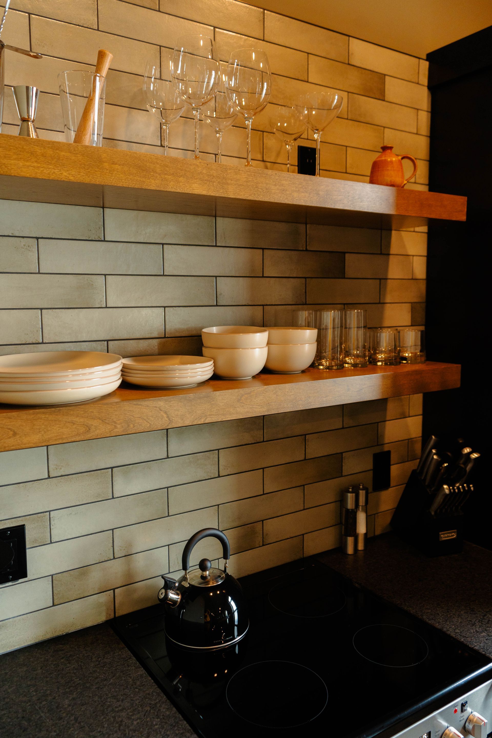 Modern kitchen with wooden shelves, stacked dishes, glassware, and a black stovetop with kettle.