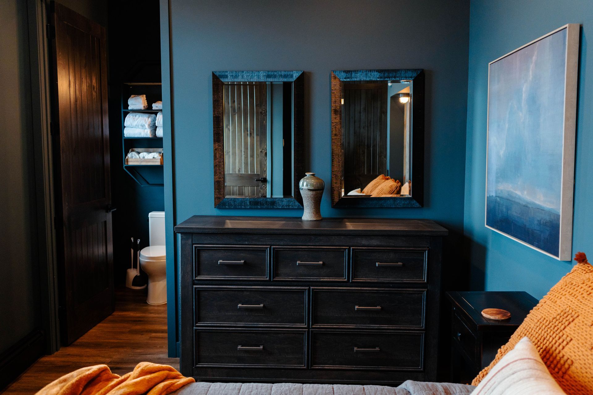 Bedroom with dark dresser, mirror, blue walls, and orange bedspread in soft light
