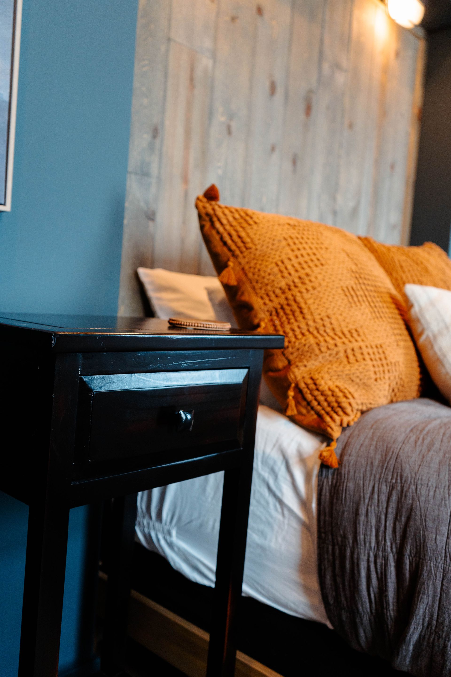 Cozy bedroom corner with a bed, orange pillow, gray blanket, and black bedside table next to blue and wood walls