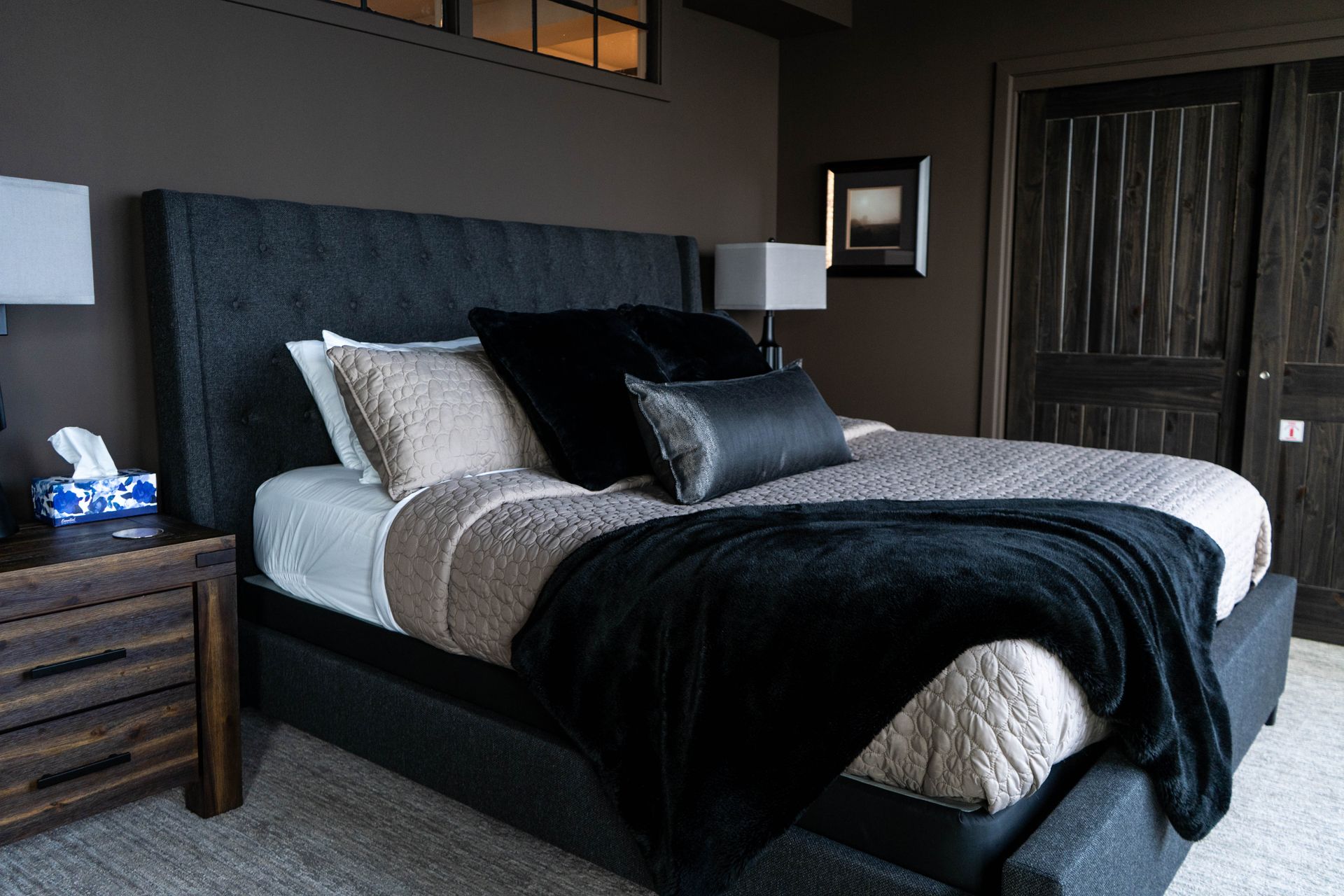 Modern bedroom with a dark upholstered bed, layered gray and black bedding, nightstands, lamp, and closet doors.