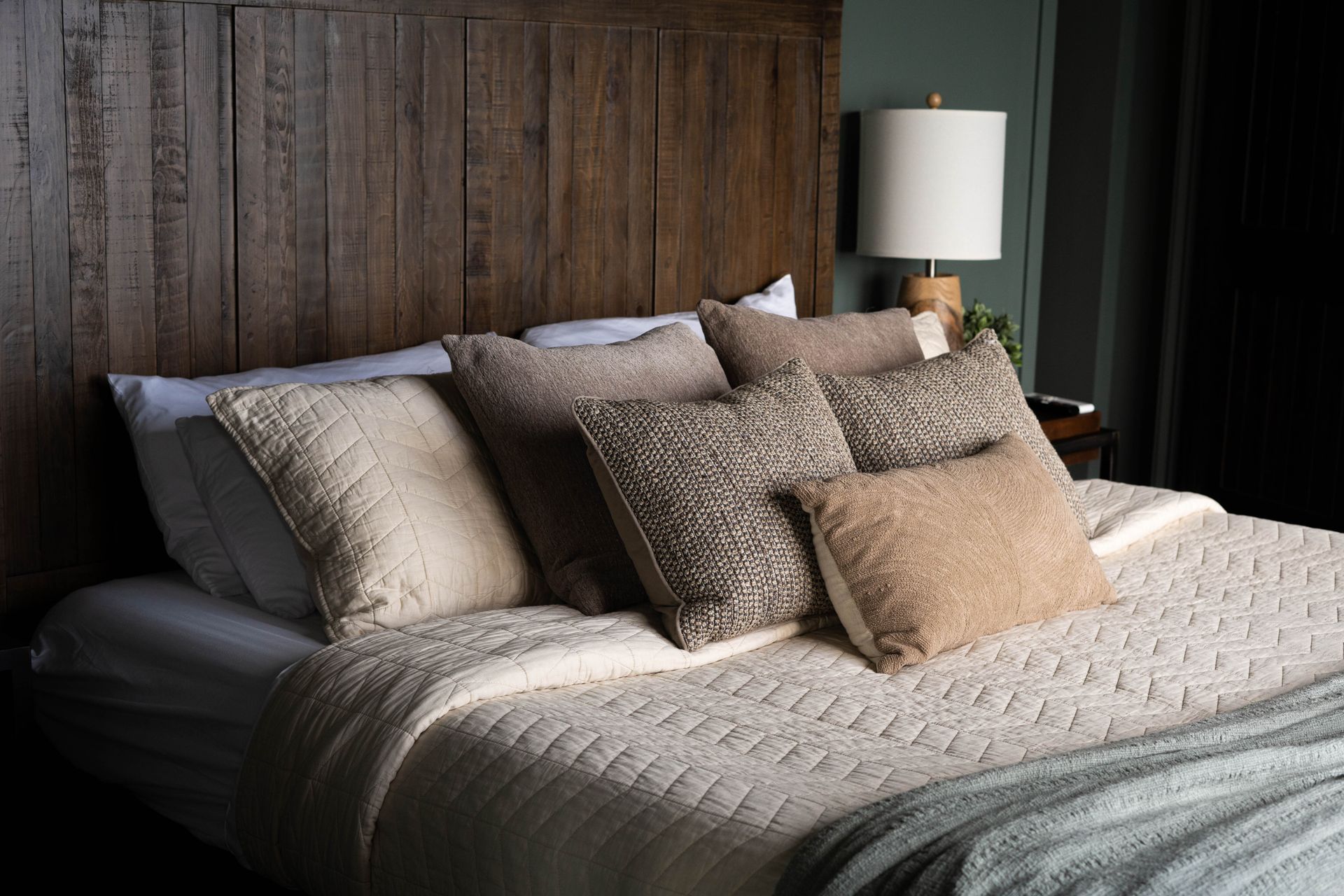 Cozy bed with layered beige pillows and textured bedding against a dark wood wall under a bedside lamp
