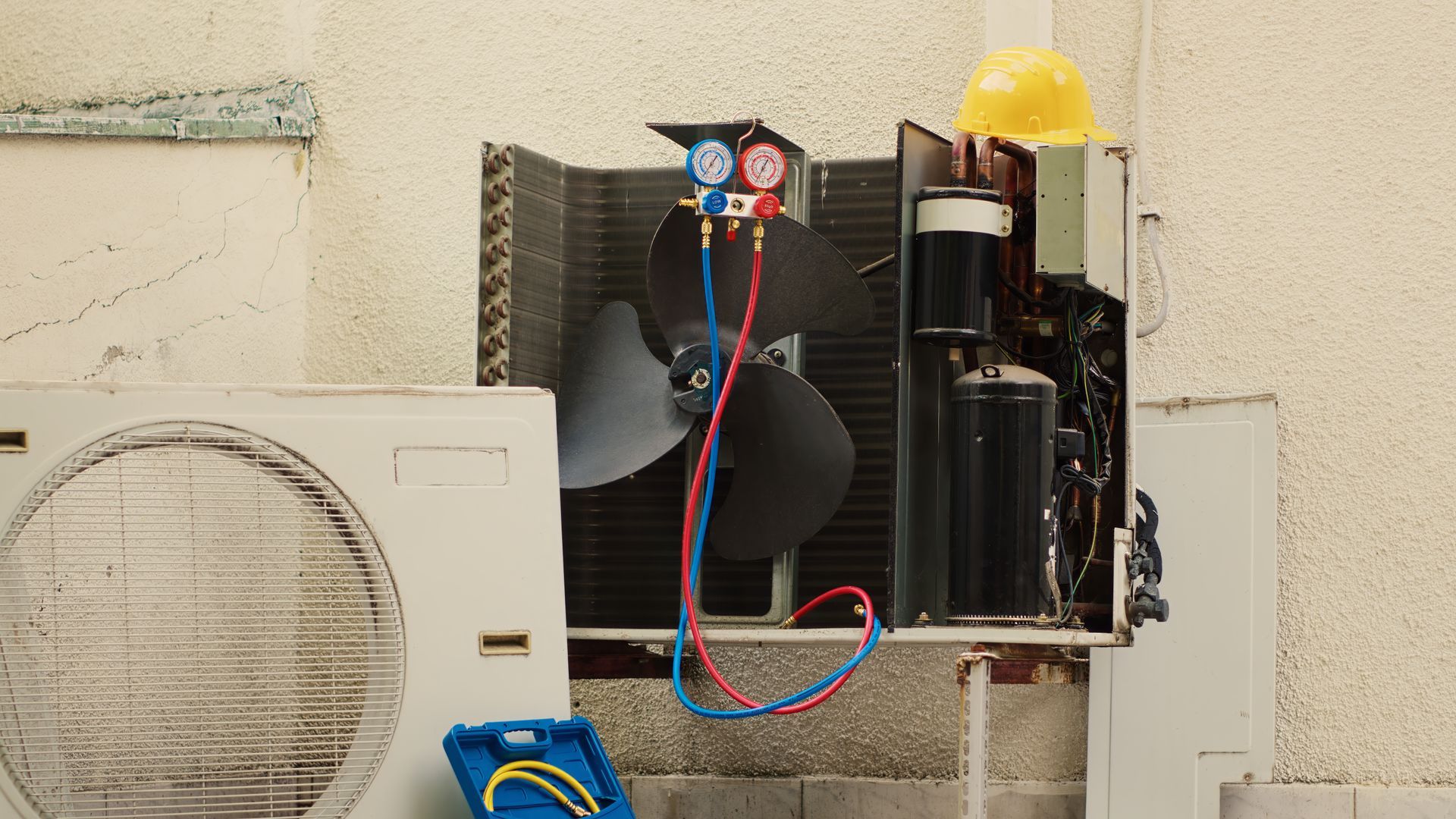 Outdoor wall-mounted mechanical unit with exposed wiring and a yellow hard hat on top, beside an AC unit.