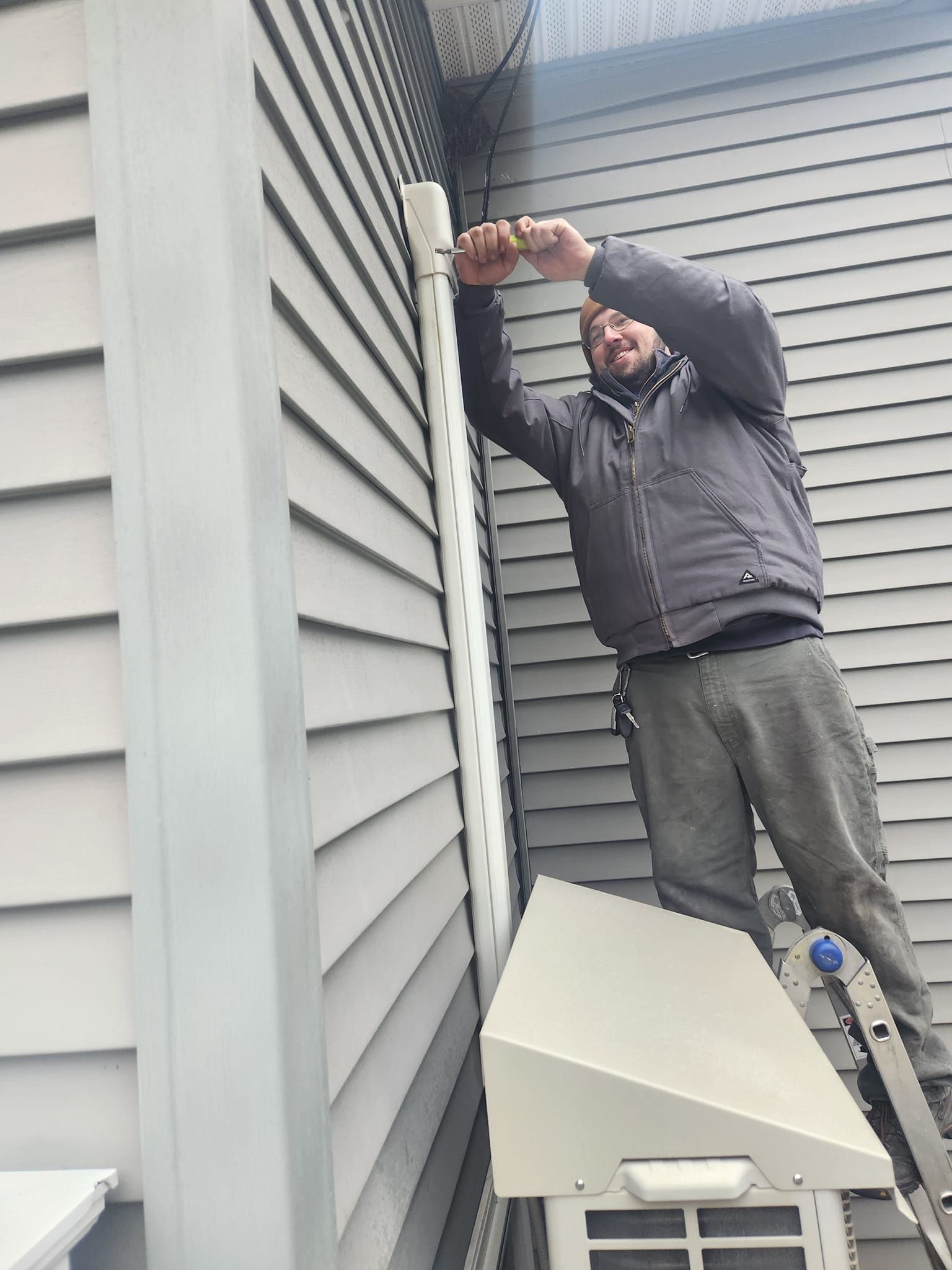 Man tightening an outdoor wall-mounted device beside a house with siding and a snow-covered roof.