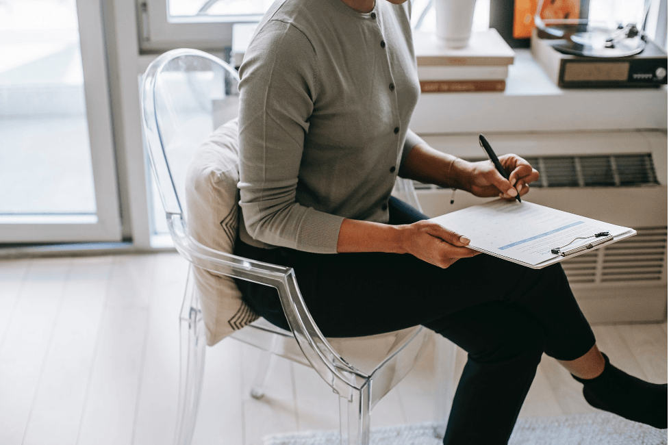 A woman is sitting in a clear chair holding a clipboard and a pen.