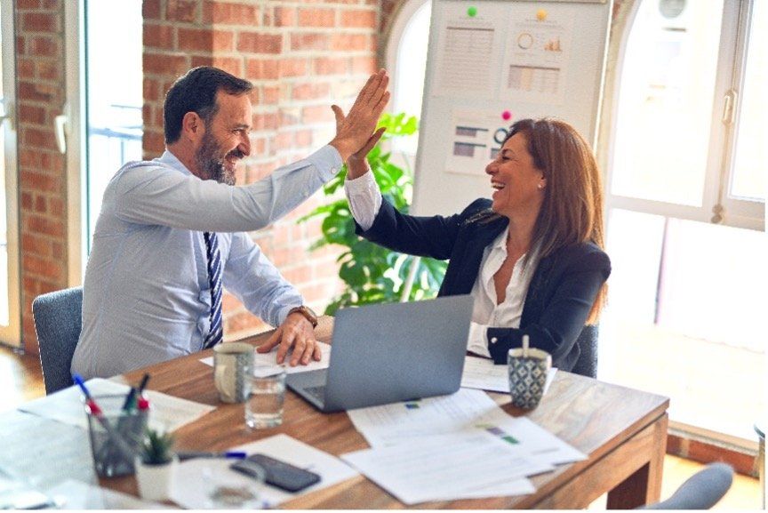 A man and a woman are giving each other a high five while sitting at a table with a laptop.