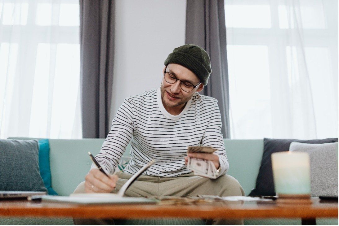 A man is sitting on a couch writing in a notebook while holding a cell phone.