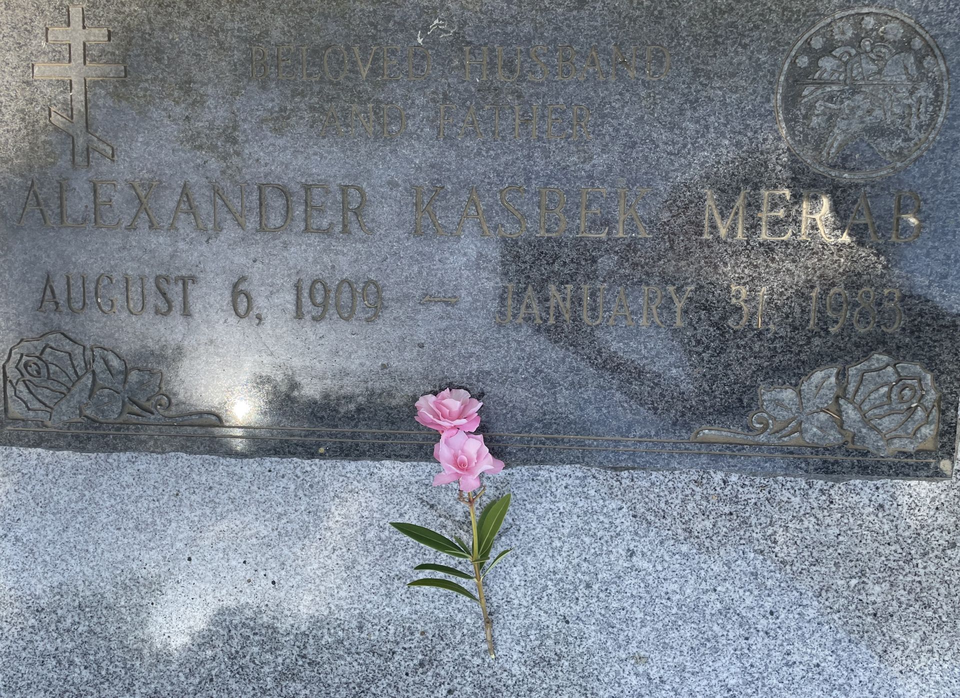 Gravestone with engraved name and dates, decorated with a pink flower in front.