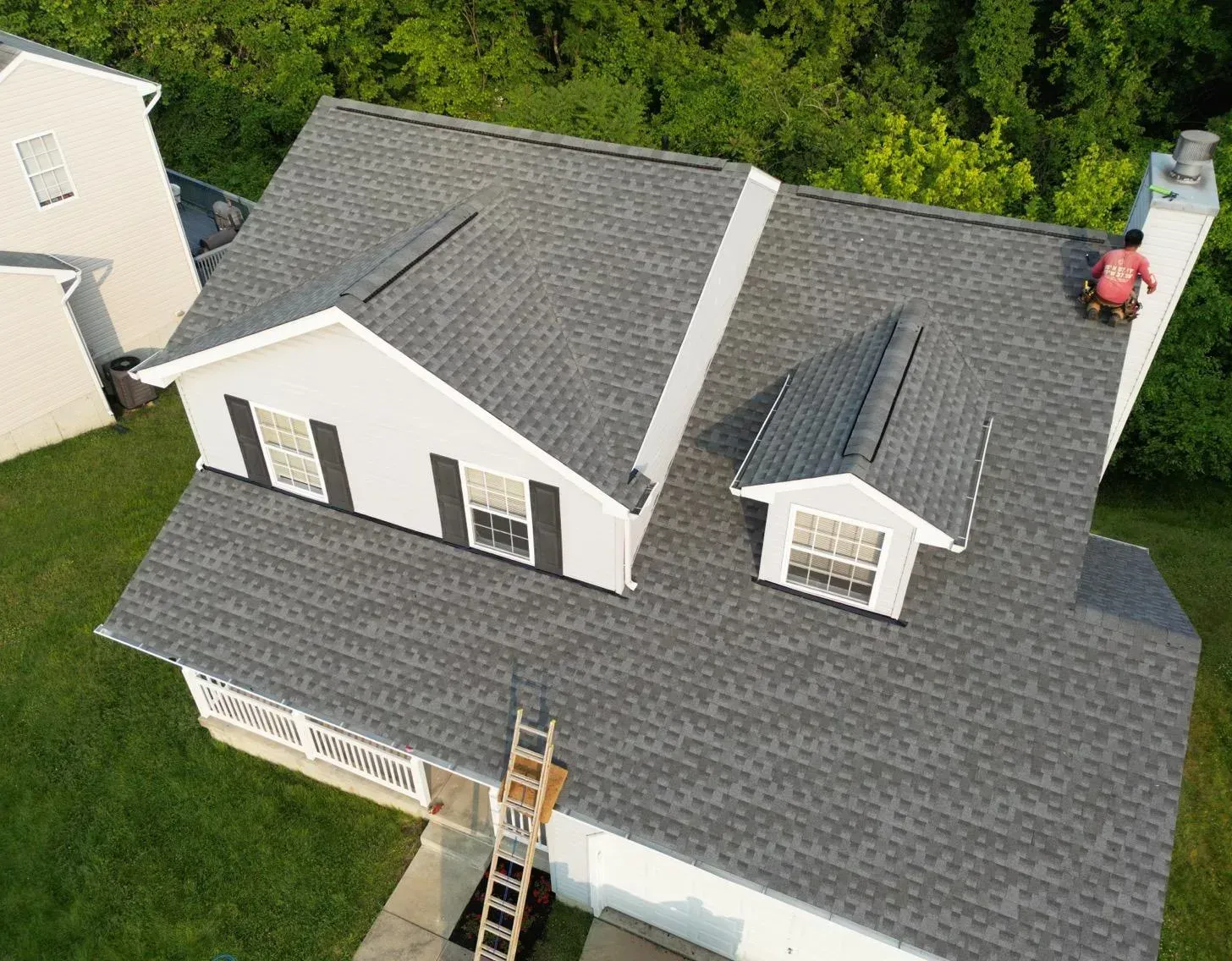 Aerial view of a gray-roofed house with a ladder on the front roof and green trees around it.