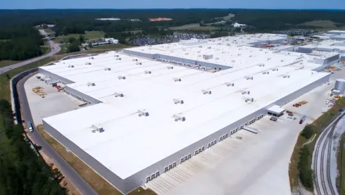Aerial view of a large white warehouse complex with surrounding roads and forested land.