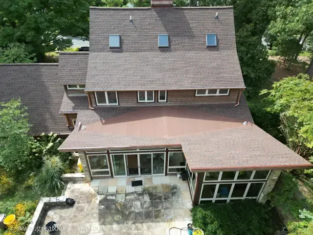 Aerial view of a large brown house with a sloped roof, skylights, and a patio surrounded by trees.