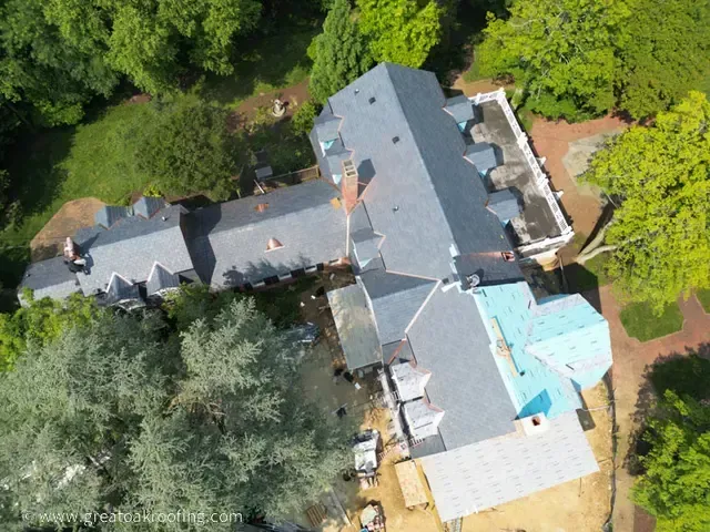 Aerial view of a large gray-roofed house surrounded by dense green trees and a small blue-roofed section.