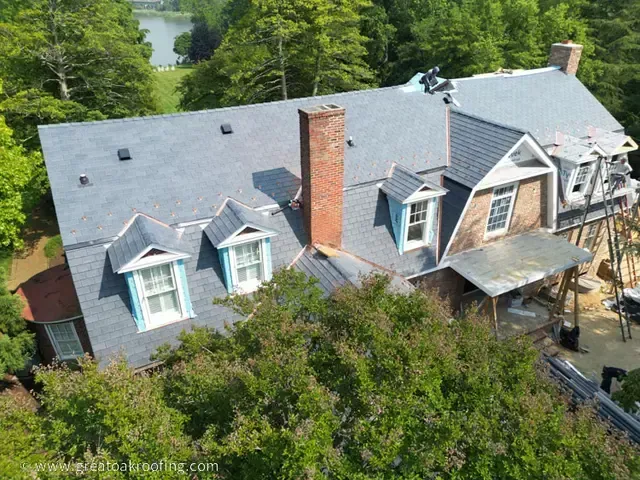 Aerial view of a gray-roofed house with dormer windows and brick chimney surrounded by trees