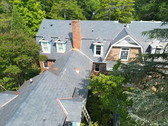 Aerial view of a large gray-roofed house with chimneys surrounded by green trees