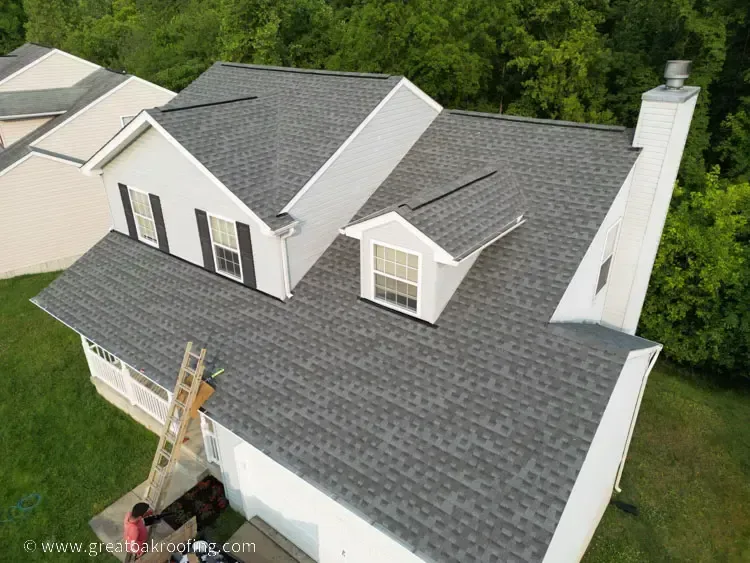 Aerial view of a house with gray shingle roof and a ladder leaning against the side amid trees.