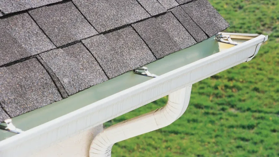 White gutter with leaf guard on house roof, collecting runoff beside gray shingles.