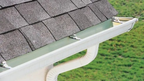 White gutter with leaf guard on house roof, collecting runoff beside gray shingles.
